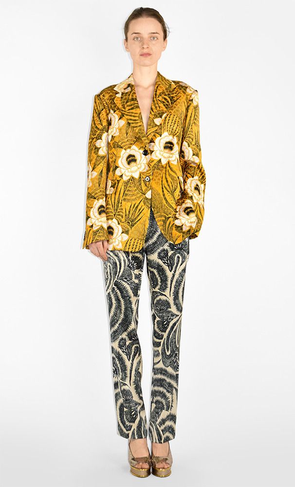 a fullbody studio shot of a woman wearing a yellow floral blazer and black and beige patterned trousers. she is standing and wearing gold sandals. the background is plain white.
