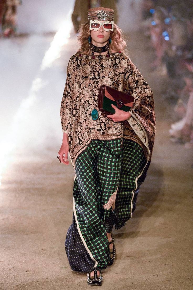 a model walks the runway at a gucci fashion show. she is wearing a unique and stylish outfit that combines several elements. the outfit features a long, loosefitting, patterned kaftan or poncho with floral and paisley designs in shades of gold, beige, and brown.  underneath, she sports widelegged palazzo pants in a dark green and black geometric pattern. the pants have a subtle color variation creating a unique effect.  she accessorizes with a jeweled headpiece, oversized sunglasses with a distinctive frame, a darkcolored, possibly leather, clutch bag, and layered necklaces. the overall style blends bohemian and vintage elements with a modern highfashion twist.  the setting appears to be outdoors, with some atmospheric effects, suggesting a themed runway show. the model's footwear seems to be darkcolored sandals with a detail near the toes.