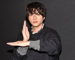 a black and white headshot of a young asian man with short, dark hair. he is making a gesture with his hands, his left hand open and his right hand touching his left wrist. he is wearing a dark gray collared shirt. the background is black.
