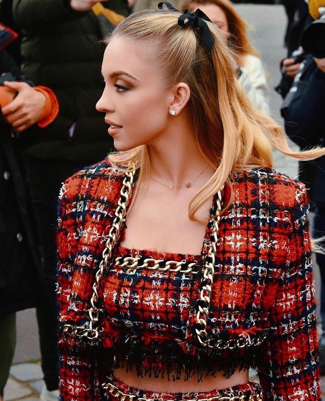 closeup shot of a blonde woman with her hair in a high ponytail adorned with a black bow. she's wearing a stylish red and black plaid tweed jacket with a gold chain detail. the woman appears to be at a fashion event, possibly a street style photoshoot. her makeup is subtly done, emphasizing her features, and she wears small earrings and a delicate necklace. the overall impression is one of high fashion and elegance.