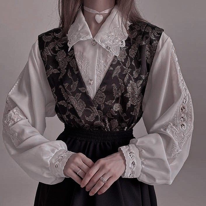 a person wearing a white blouse with lace detailing and a black floral vest, paired with a black skirt. they are also wearing a white choker with a heart pendant and have rings on their fingers.