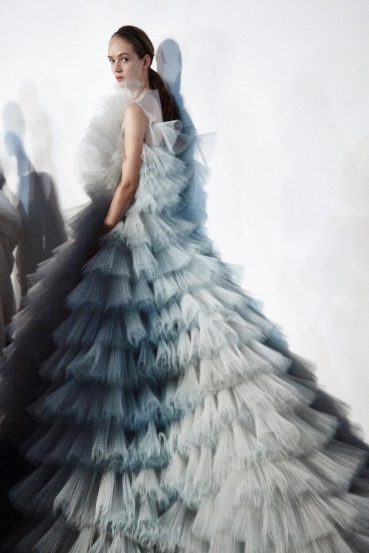 a woman in a stunning blue and white tulle gown with numerous ruffles poses for a photoshoot. the gown is ethereal and dreamlike, and the woman's pose is both elegant and glamorous. the background is a simple white wall, which allows the dress to be the focus of the image.