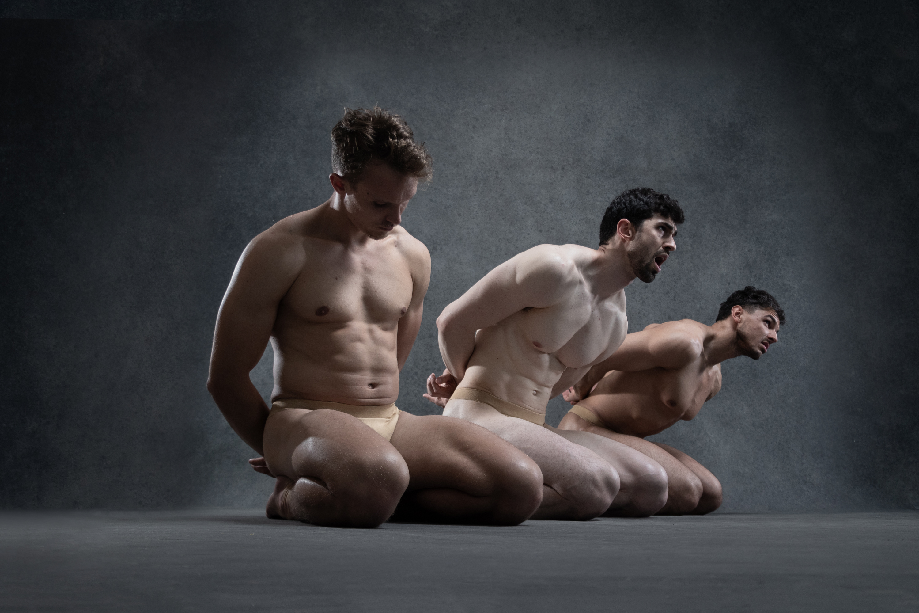 three muscular male dancers are captured midpose against a dark, textured background. they are shirtless and wear fleshtoned dance briefs, their bodies illuminated by dramatic studio lighting.  all three are crouched low, arms pulled behind their backs, their faces turned to the right in a progression of intense expressions. the overall feeling is one of dynamic movement frozen in time, highlighting the strength and emotionality of the dancers and their performance.