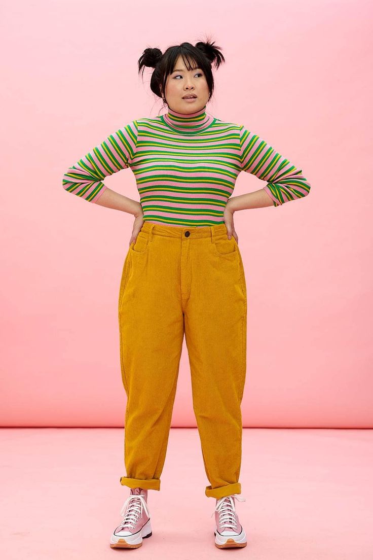 a fullbody shot of a young woman against a solid pink background. she's wearing a pink and green striped longsleeved turtleneck top tucked into highwaisted mustard yellow corduroy pants. her hair is styled in two top buns. she's wearing pale pink converse sneakers. she is standing with her hands on her hips and has a slightly upward gaze, giving a relaxed and happy expression. the overall style is casual, reminiscent of 90s or retro fashion.