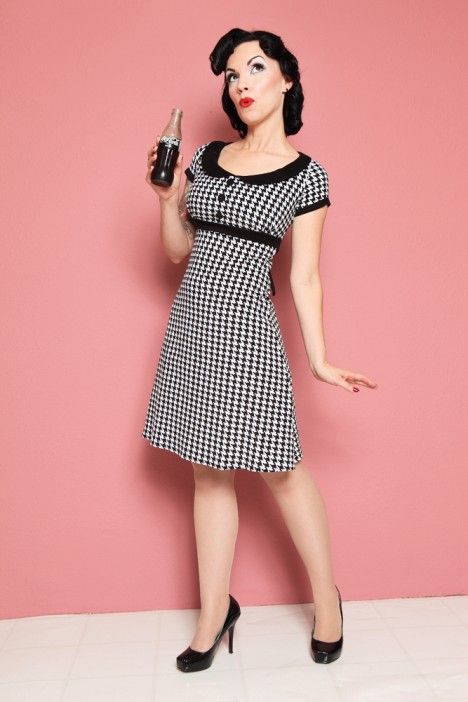 a woman is standing in a pinup pose. she is wearing a black and white houndstooth dress with a peter pan collar and black trim. she has black hair styled in a vintage fashion, and is wearing black high heels. she is holding a bottle of cocacola in her right hand, and is looking up and to the left. the background is a solid pink color.