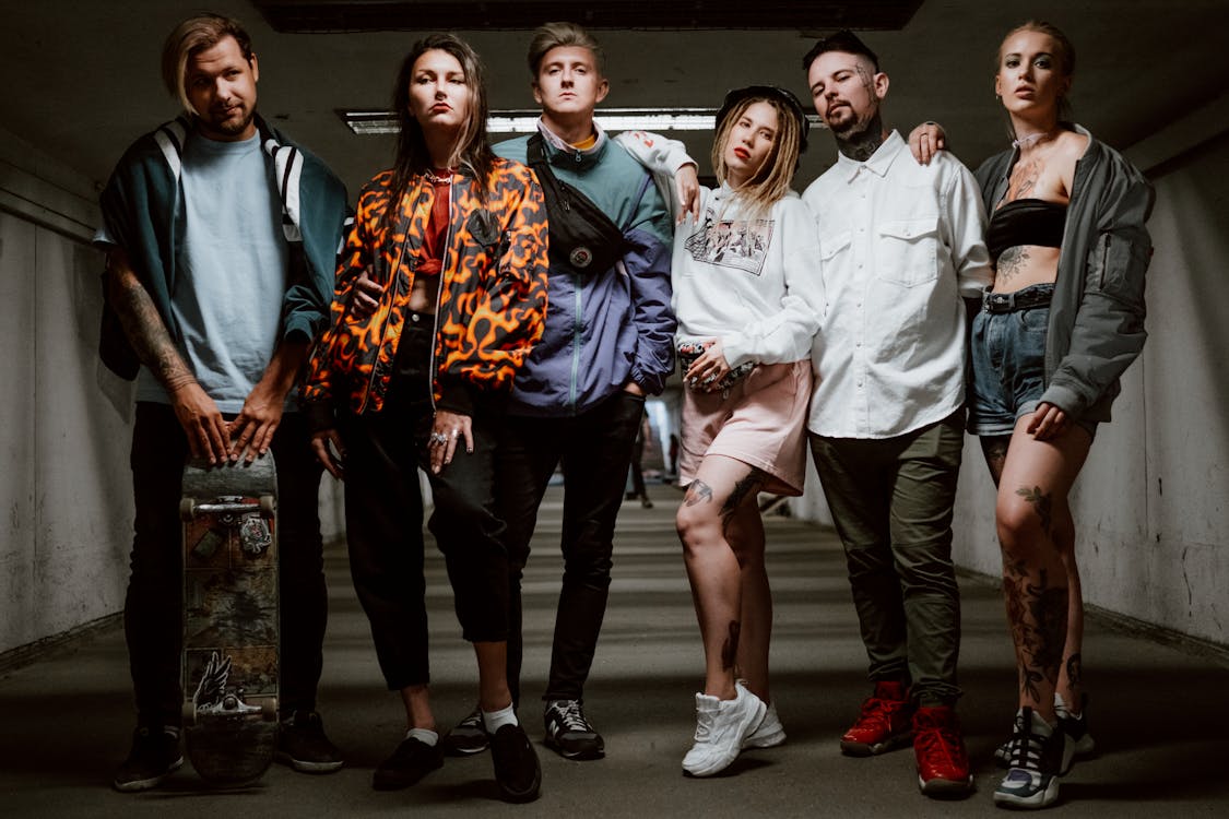 the image shows a group of six young adults, three men and three women, standing in an urban underpass.  they are all dressed in stylish and somewhat alternative clothing.  the styles range from streetwearinspired looks to more grungeleaning attire. several group members have visible tattoos. one of the men holds a skateboard.  the overall mood is cool, confident, and slightly rebellious.  the setting and poses emphasize a sense of urban youth culture. the lighting is moody and dramatic.