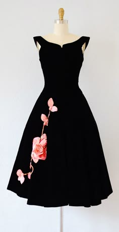 a black vintage dress with a pink flower design on the side is displayed on a white background. the dress has a square neckline and is made of a soft, flowing fabric. it is a classic and elegant piece that would be perfect for a special occasion.