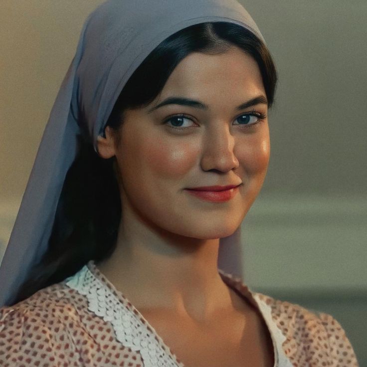 the image shows a closeup portrait of a young woman with dark hair and fair skin. she is wearing a light grey headscarf or hijab that covers most of her hair and flows down her back. she is smiling gently, with a serene and peaceful expression on her face.  her clothing consists of a light brown, polkadot dress with a white lace collar. the background is muted and slightly out of focus, and the lighting is soft and warm, giving the image a vintage or antique feel. the overall mood is calm and elegant, suggesting a possible film still or historical setting.