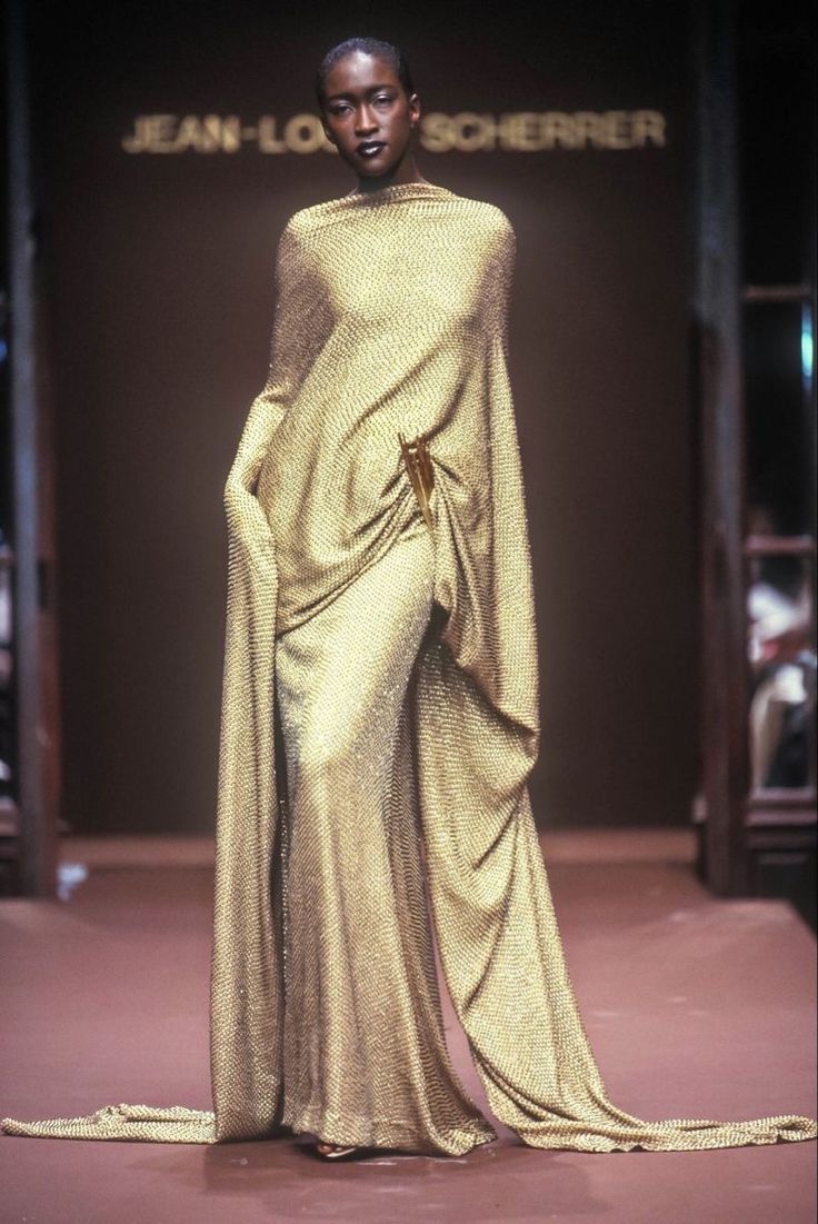 a black female model walks the runway in a shimmering gold gown, her pose is poised and elegant. the gown is long and flowing, and the fabric seems to be metallic, creating a striking look. the runway is lined with a brown surface. the background features a brown wall with a banner saying "jeanlouis scherrer" in white text.  the model is walking towards the camera.