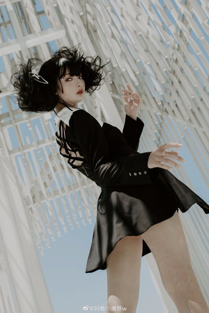 a woman with black curly pigtails and bangs is wearing a black and white dress. she is standing in front of an architectural background with a light blue sky visible. she is looking at the camera with her fingers pointed. she has jewelry on her fingers, including a ring. the dress has buttons and a skirt that flares out.