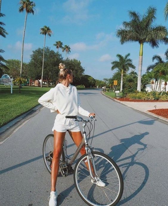 a woman is standing in the street with her bicycle on a sunny day. she is wearing a white sweatshirt and shorts, with her hair in a bun. there are palm trees and tropical foliage in the background, and the sky is blue.