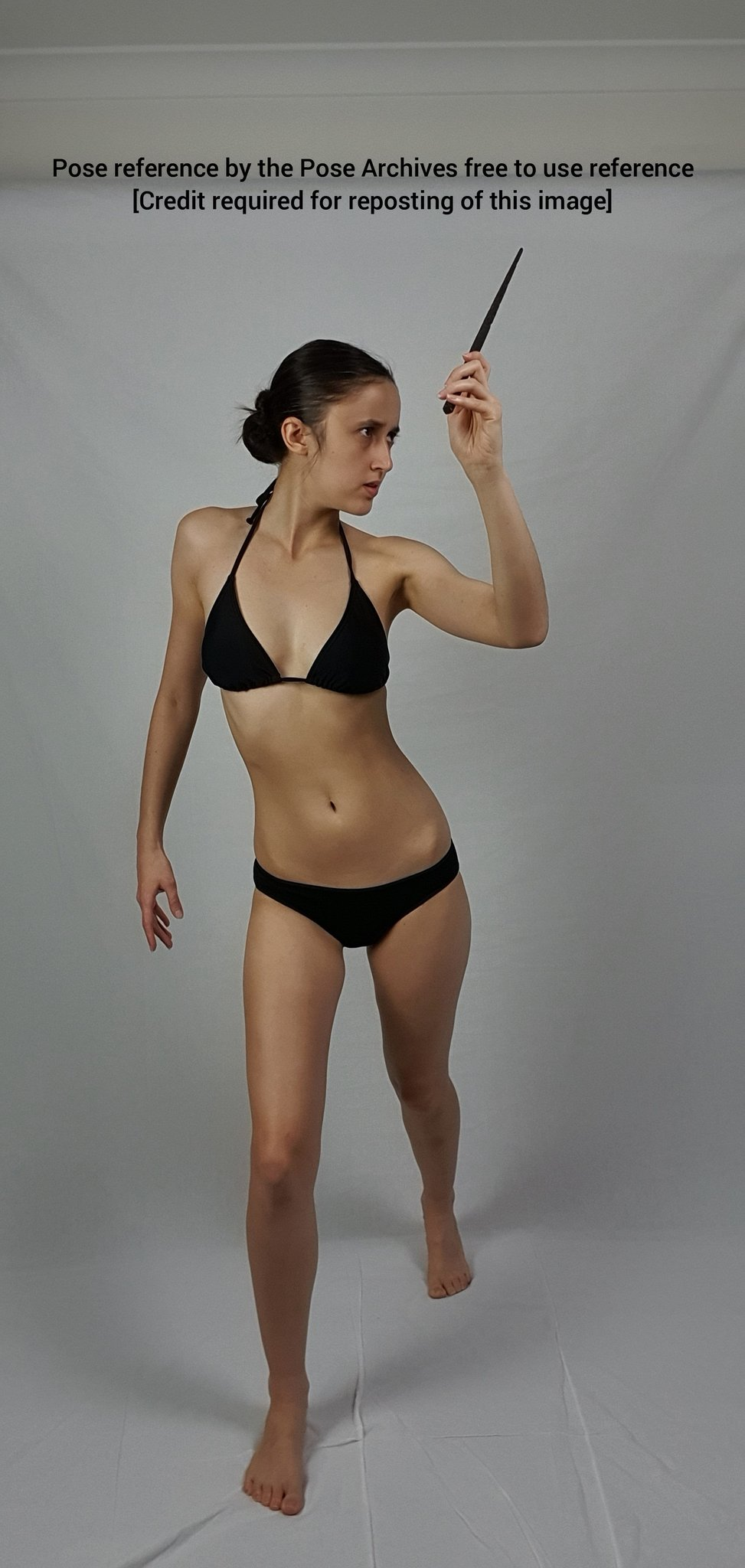 a woman in a black bikini stands against a white background, holding a wand in her right hand. she is looking at the wand. the image is a pose reference, free to use with credit.```