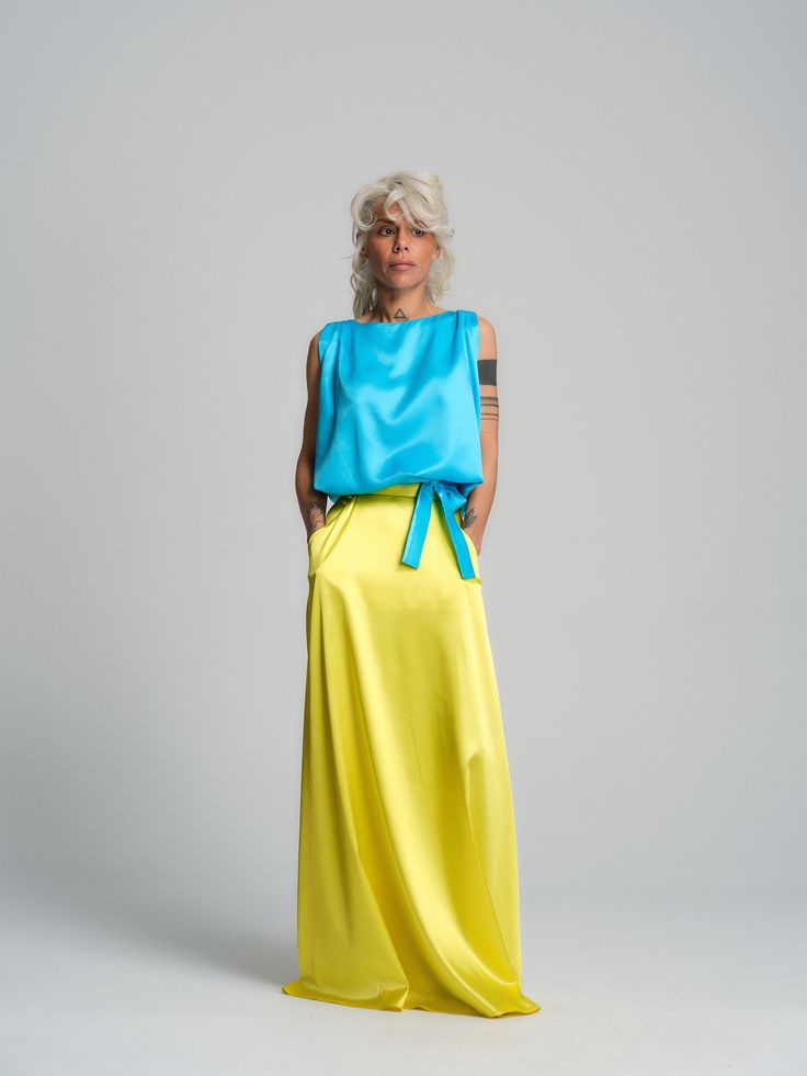 a woman with short white hair is wearing a blue silk top with a yellow silk skirt. she is posing in a studio, with a white background.