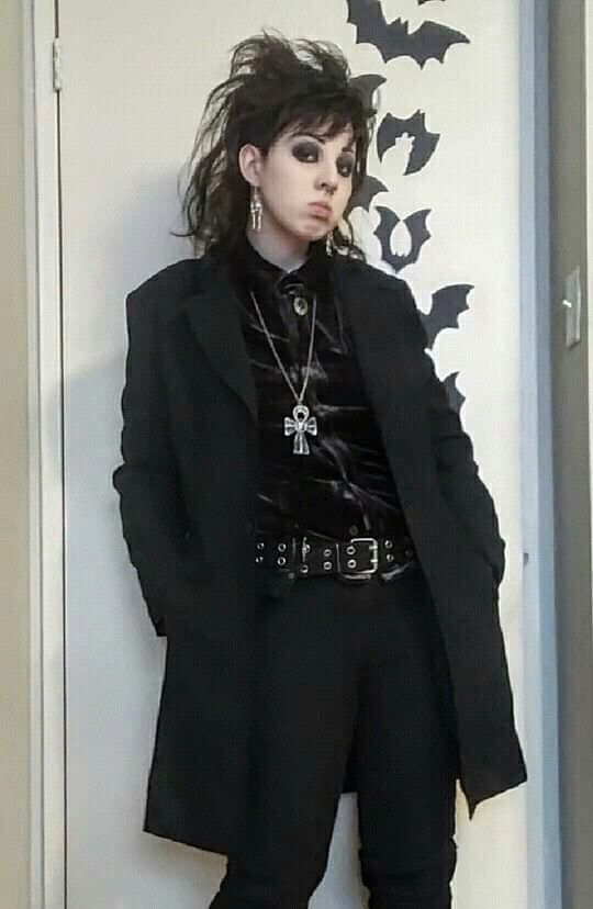 a woman in a gothic fashion style with dark hair, eyes, and a black coat, choker, and necklace stands with her hands tucked into her pockets. she is standing in front of a wall decorated with bat silhouettes.