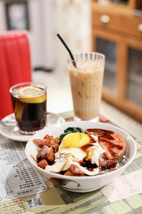 a closeup shot of a bowl of food, typically found in a hong kongstyle cafe (cha chaan teng). the bowl contains a fried egg with a runny yolk, char siu (barbecued pork), luncheon meat, rice, and spinach.  a dark brown sauce is drizzled over the dish.  in the background, there are glasses of iced milk tea and a colalike drink with a lemon slice. a menu is visible on the table. the setting appears casual and comfortable, suggesting a local eatery.