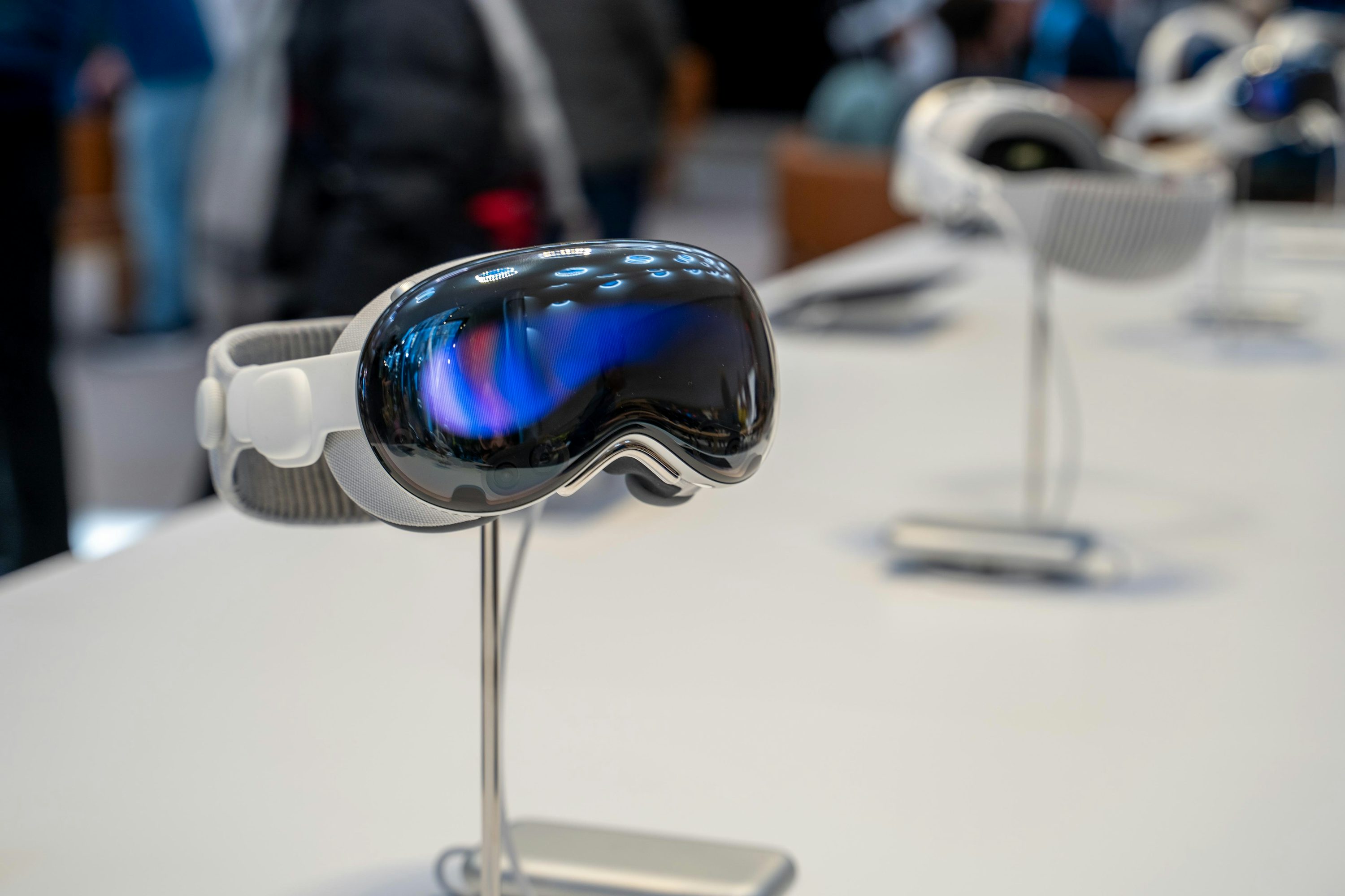 a closeup shot of an ar headset on a silver stand. the headset has a silver frame and a dark reflective display. several more headsets can be seen in the background.