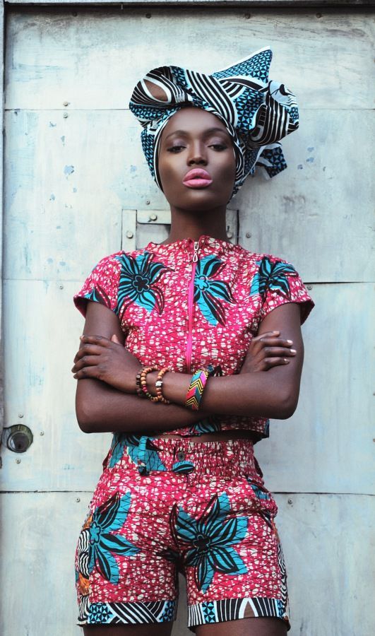 a young woman wearing a vibrant africaninspired outfit, featuring a patterned headwrap and a matching crop top and shorts. she stands confidently against a grey wall, her expression bold and cool.