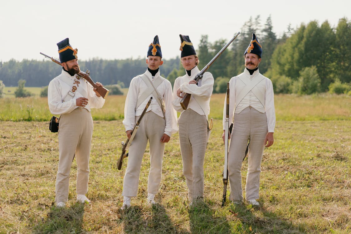 four men dressed in authentic 18thcentury military uniforms stand in a grassy field, holding muskets. they are likely participating in a historical reenactment.  the uniforms are light beige with white waistcoats, and the men wear tall, dark hats. the scene evokes a historical military setting.