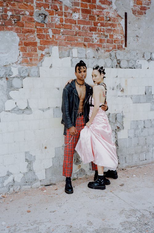 a young couple stands against a weathered brick wall. the man is wearing a black leather jacket, red plaid pants, and black boots. the woman wears a pink satin dress and black boots. they are embracing, showcasing a punkinspired fashion style. the overall mood is edgy and rebellious. the urban backdrop adds to the scene's grittiness.  the image suggests a photoshoot showcasing alternative fashion.