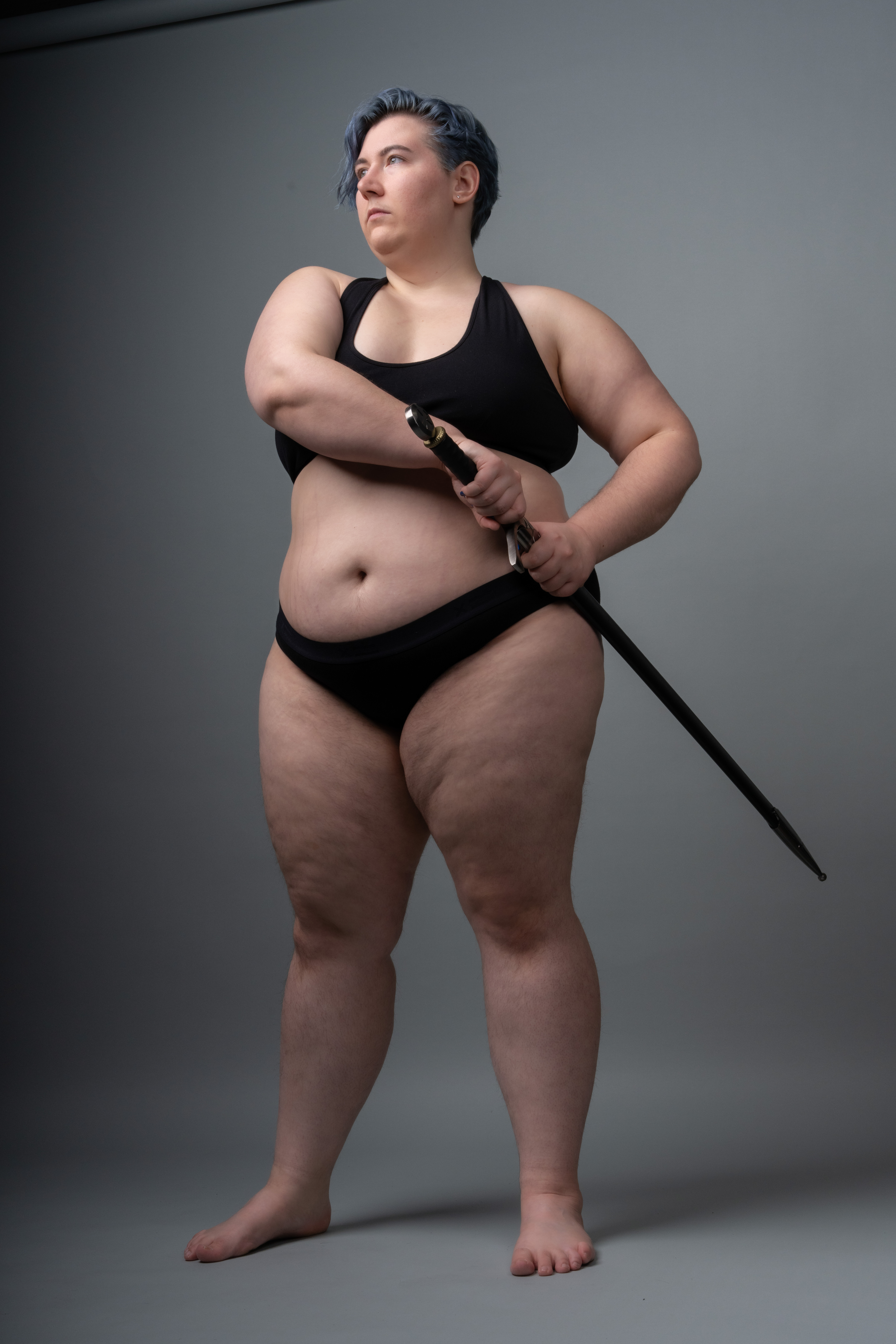 a plussize woman with blue hair is standing barefoot in a studio with a grey background. she is wearing a black bra and black underwear and holding a sword in front of her. she is looking away with a serious expression.