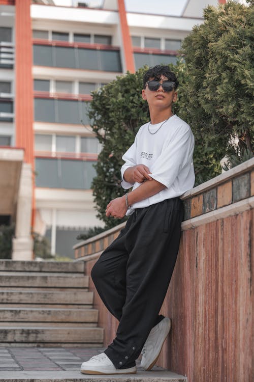 a young man is posing for a photo outdoors, leaning against a wall with a set of stairs to his left. he is wearing sunglasses, a white tshirt, black pants, and white sneakers. he is standing in front of a building with a tree behind him.