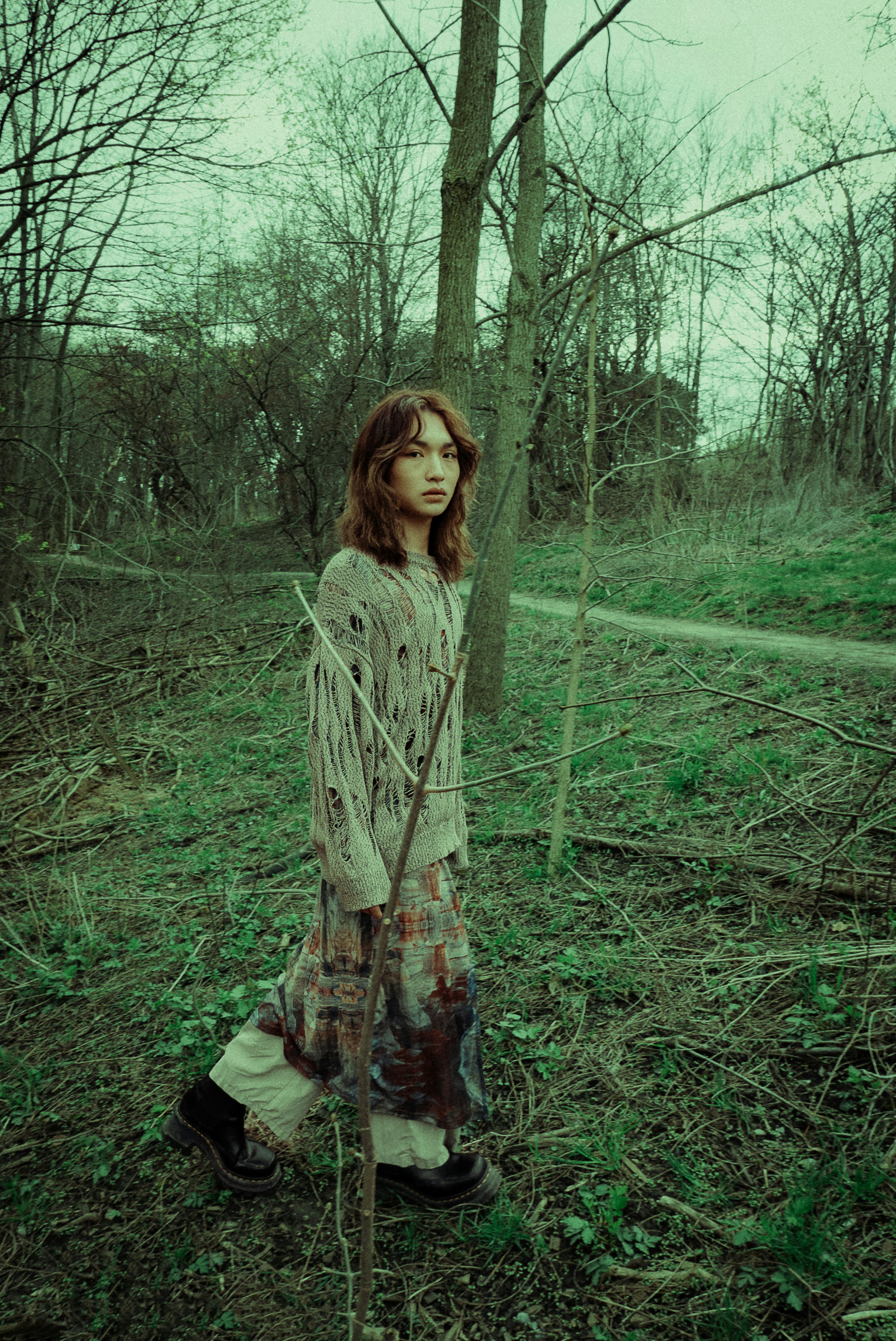 a woman stands in a forest, wearing a distressed sweater, a patterned skirt, and black boots. she is surrounded by trees and green foliage. a path is visible in the background under an overcast sky.