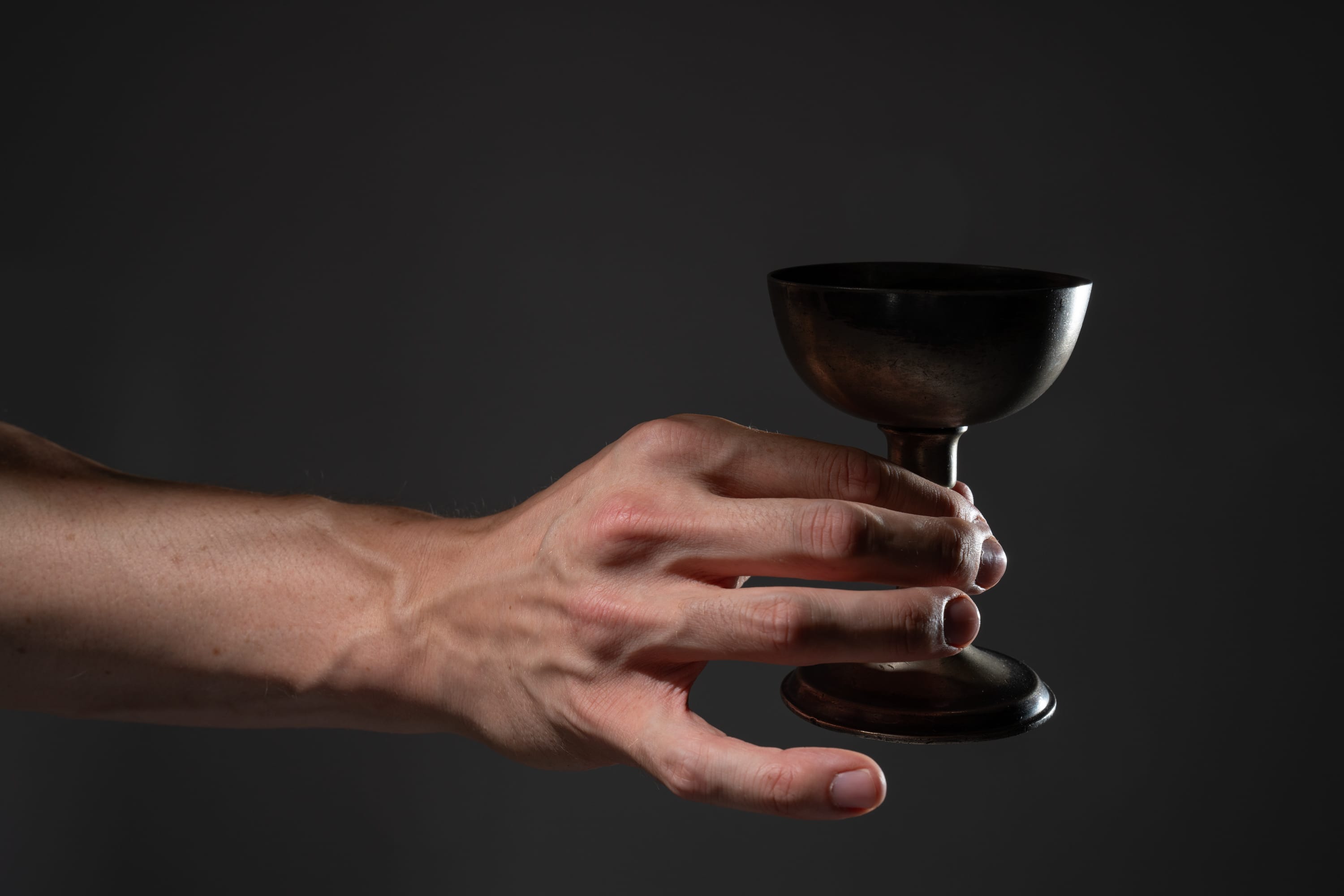 a man's hand, illuminated by a dramatic spotlight, holds an antique, metallic chalice against a dark background. the lighting emphasizes the texture of the hand and the cup, creating a sense of weight and significance.