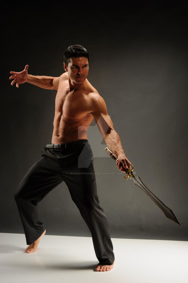 a muscular man in black pants is shown standing on a grey background with a sword in his right hand, he is shirtless, his left hand is raised in a gesture that appears to be calling out in the direction of his left hand. the man is looking in the direction of his left hand. there are shadows created by the lighting in the studio.