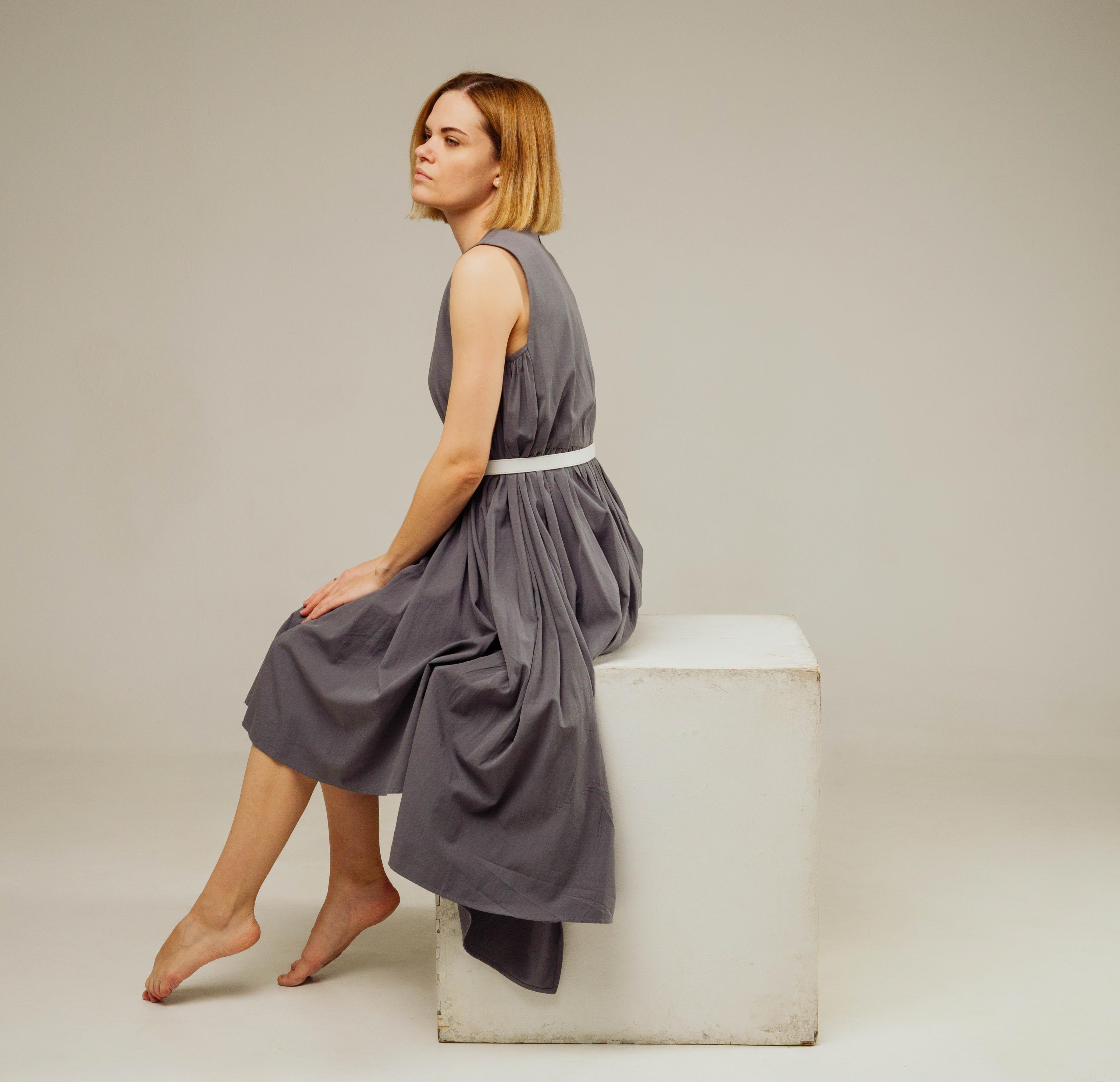 a woman in a gray dress is sitting on a white cube. she is looking to the left of the frame with a neutral expression. the background is a light gray.