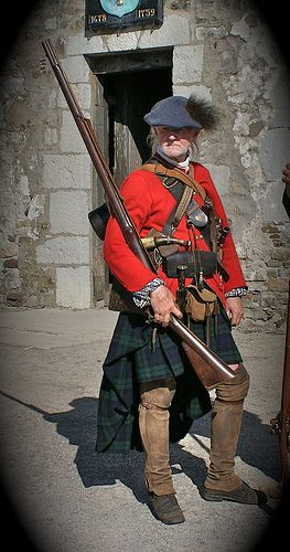 a man in a historical reenactment outfit stands holding a musket. he wears a red coat, kilt, hat, and boots, typical of a scottish soldier in the 17th or 18th century. he stands in front of a stone building, possibly a historical site.