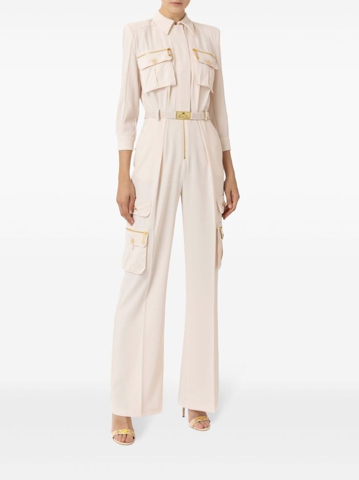 a woman is wearing a white jumpsuit with long sleeves, a gold belt, and zipper detailing. it has pockets and a loose fit. she is wearing matching high heels and is standing in a studio with a white background.