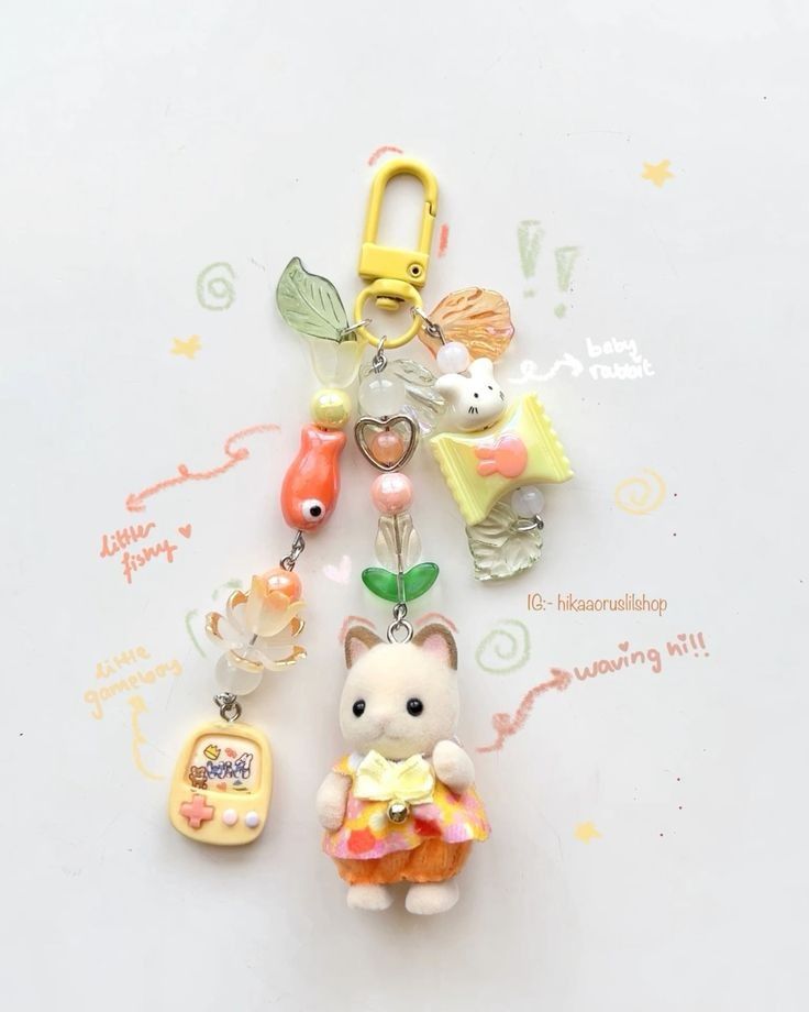 a closeup of a keychain with various charms attached. the keychain has a yellow clip at the top, followed by a chain of beads and charms including leaves, a fish, a video game console, and a rabbit. the main charm is a rabbit figurine wearing a dress. the background is white.