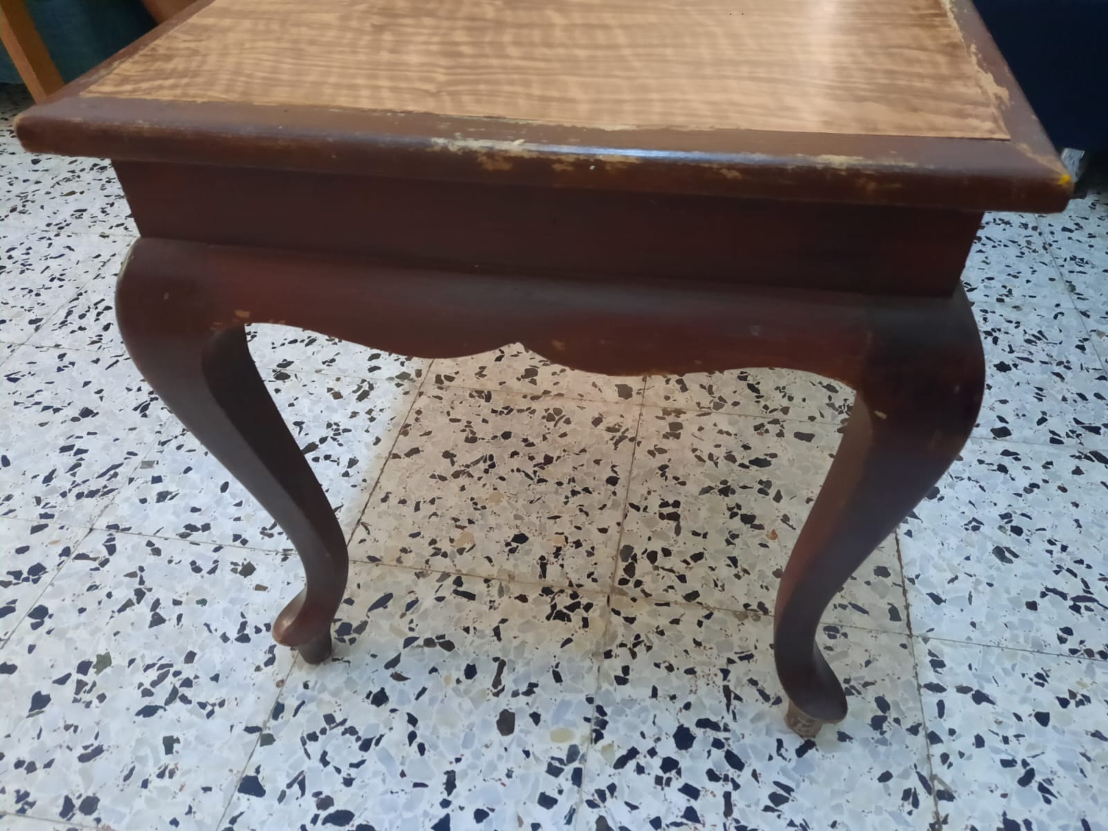 the image shows an antique wooden table with cabriole legs, standing on a tiled floor. the table has a brown frame with a lighter, patterned top. it appears to be old and shows signs of wear. the floor is tiled with a speckled pattern.