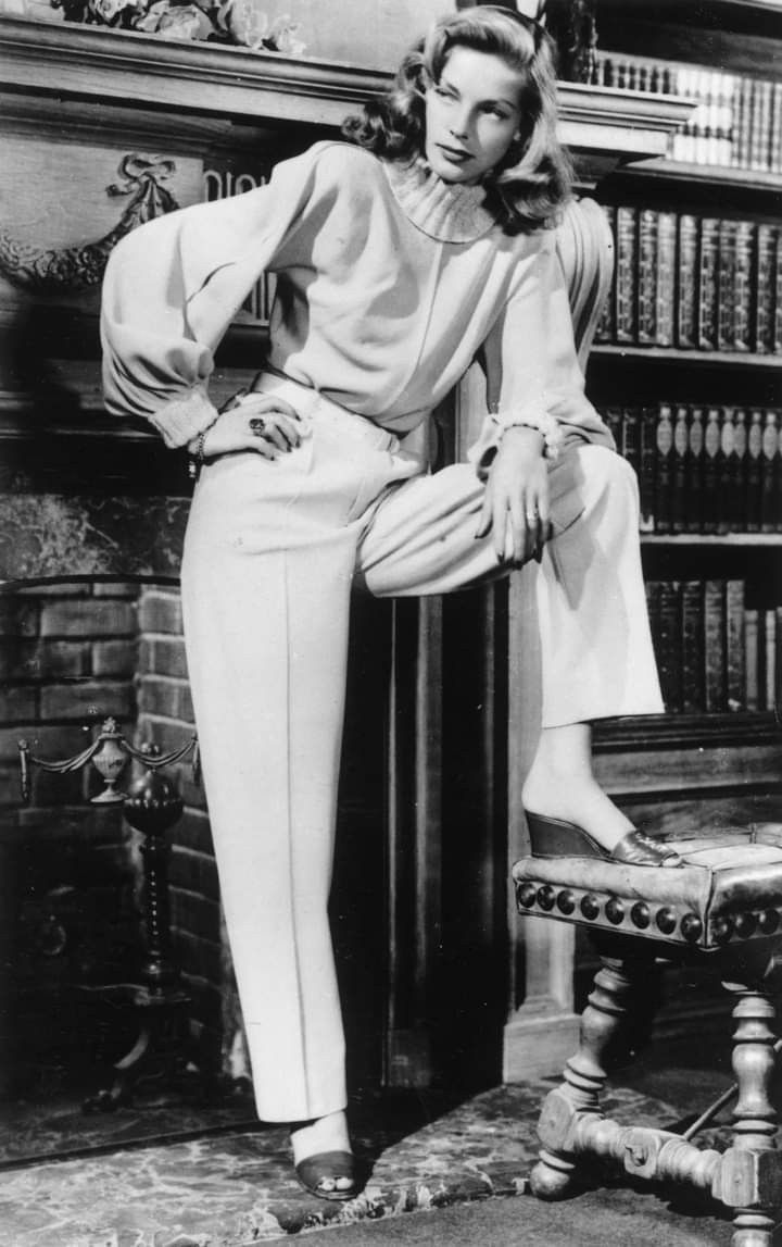 a black and white photograph of a woman in a classic 1940s or 1950s style outfit. she is wearing white pants and a white sweater. her hair is long and blonde. she is sitting on a stool with her legs crossed. she is looking off to the side and has a serious expression on her face. the photo is welllit and sharp, and the woman's features are clearly visible. the background is a bookshelf. the overall effect of the photo is glamorous and timeless. the woman could be a model or an actress.