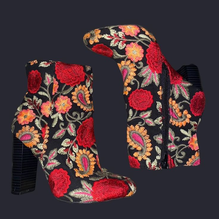 the image shows two ankle boots with black fabric and colorful floral embroidery on a dark background. the boots have a stacked block heel. the embroidery features red and orange flowers and paisley patterns. one boot is standing upright, while the other is lying on its side, revealing a zipper along the side.