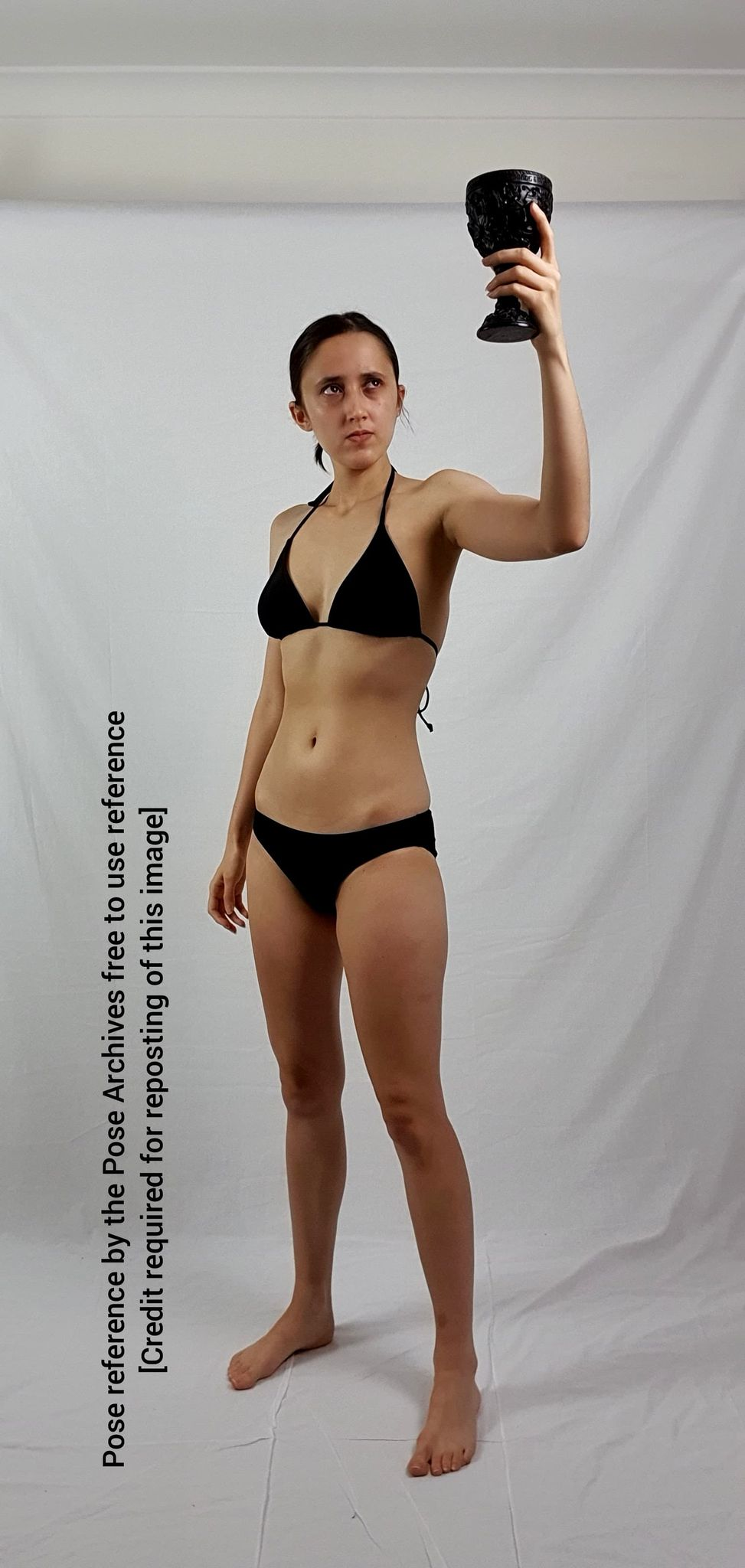 a woman in a black bikini is standing with her feet apart, holding a darkcolored goblet above her head in her right hand. she has a neutral expression on her face and appears to be posing for a photograph. the background is a plain white wall, and the lighting is soft and diffused.```