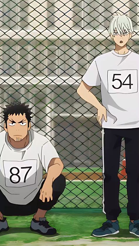 two anime boys in athletic wear stand in front of a chainlink fence. the boy on the left is crouched down, while the boy on the right stands with his hands on his hips. they are wearing numbered shirts, suggesting they are about to participate in a sporting event, potentially a track and field race.