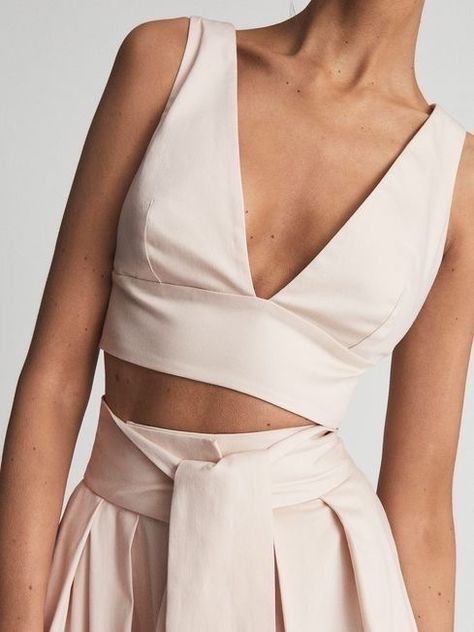 the image showcases a woman wearing a stylish summer outfit. the outfit consists of a pale pink crop top with a deep vneckline and sleeveless design. it is paired with matching highwaisted pants featuring a tiefront detail. the image is a closeup shot, focusing on the top portion of the outfit and the woman's torso. the lighting appears to be studio lighting, creating a bright and clear image.