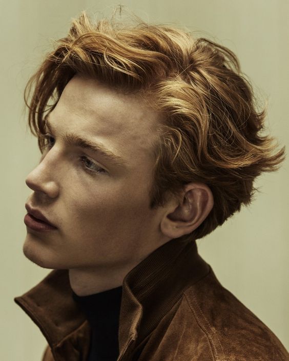 a closeup portrait of a young man with ginger hair. he is wearing a brown suede jacket and a black turtleneck. the lighting is soft and the background is blurred, creating a sense of depth and intimacy. the man's gaze is directed offcamera, giving him an air of contemplation.