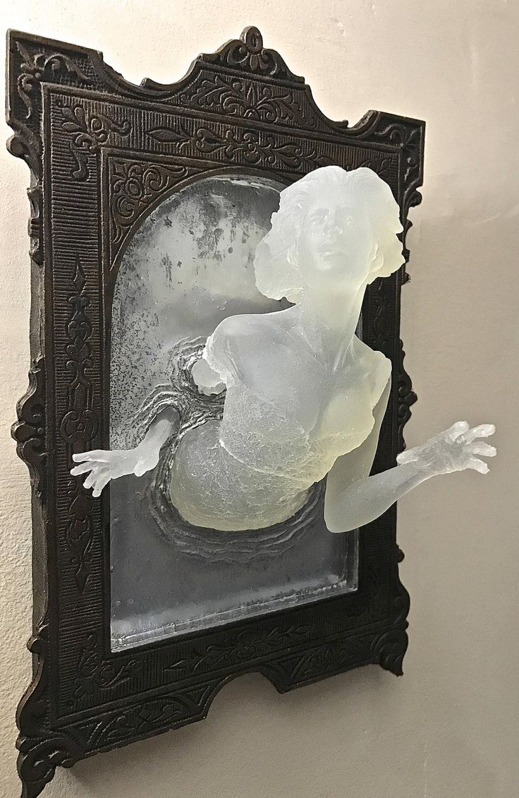 the image shows a 3dprinted sculpture of a translucent, almost ghostlike female figure emerging from an ornate, darkcolored frame. the frame resembles an antique or art deco style. the figure is partially submerged, giving the impression of it emerging from water or a mirror.  the material appears to be resin or a similar translucent material, allowing light to pass through. the overall aesthetic is both elegant and slightly eerie. the sculpture is mounted on a wall.