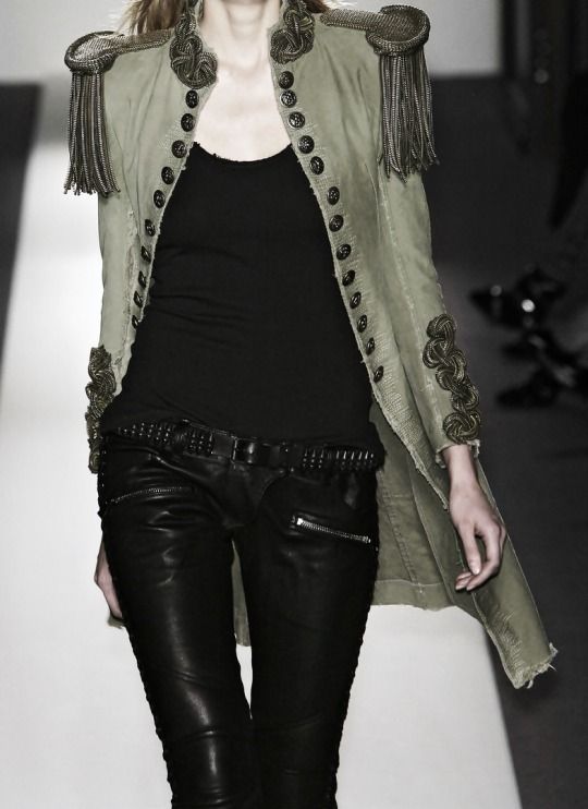 a woman on a runway is wearing a fashionable outfit consisting of black leather pants with zippers, a black top, and a militarystyle jacket with buttons, fringed shoulders, and epaulettes. a studded belt accentuates the waist.