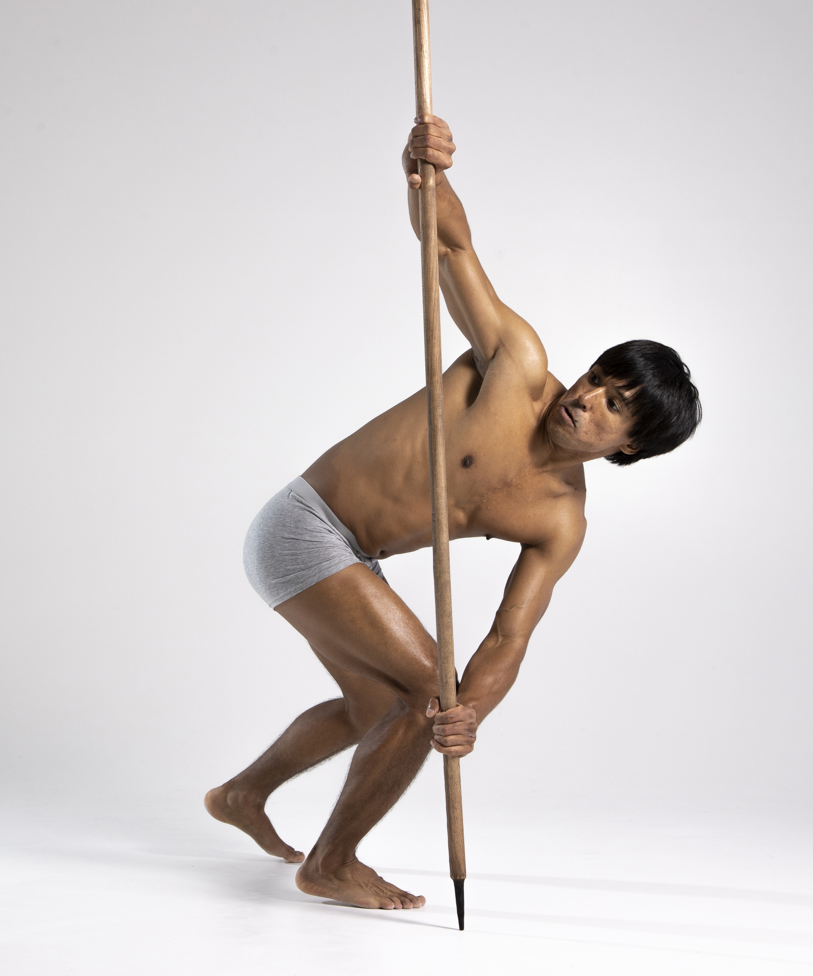 a full shot of a shirtless, muscular man wearing gray underwear and holding a long wooden spear. he is barefoot and looking at the viewer with a serious expression. the background is a plain light gray. the man is posed in a dynamic stance, leaning against the spear for support.