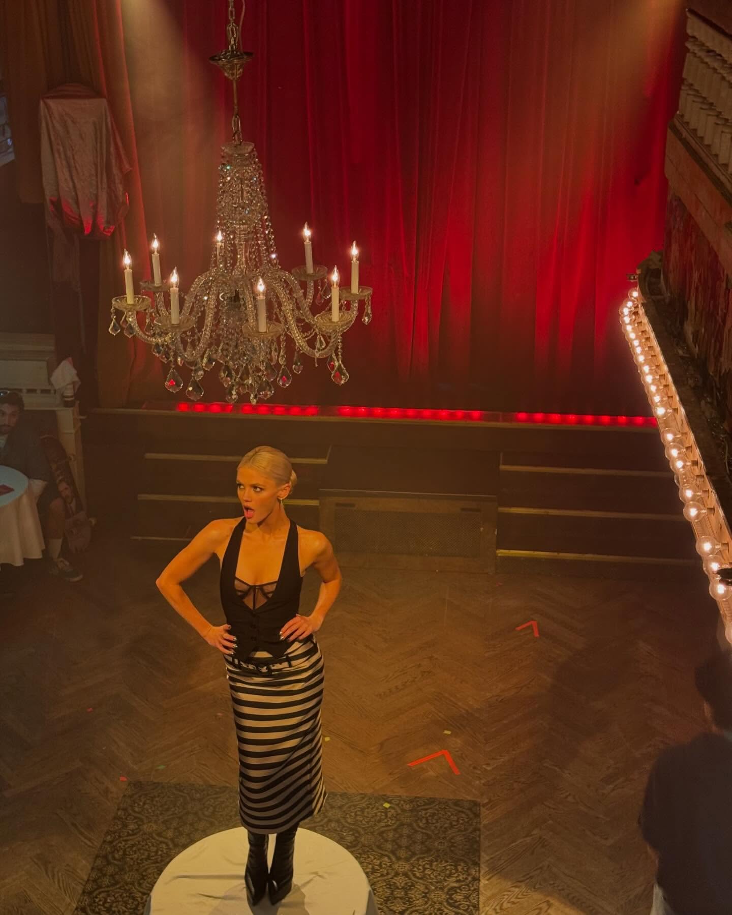 a woman in a black halter top and black and white striped skirt stands on a white round platform on a stage beneath a chandelier. behind her is a red curtain and a set of stairs. there are people seated in the background. a light is visible on the right side of the image.
