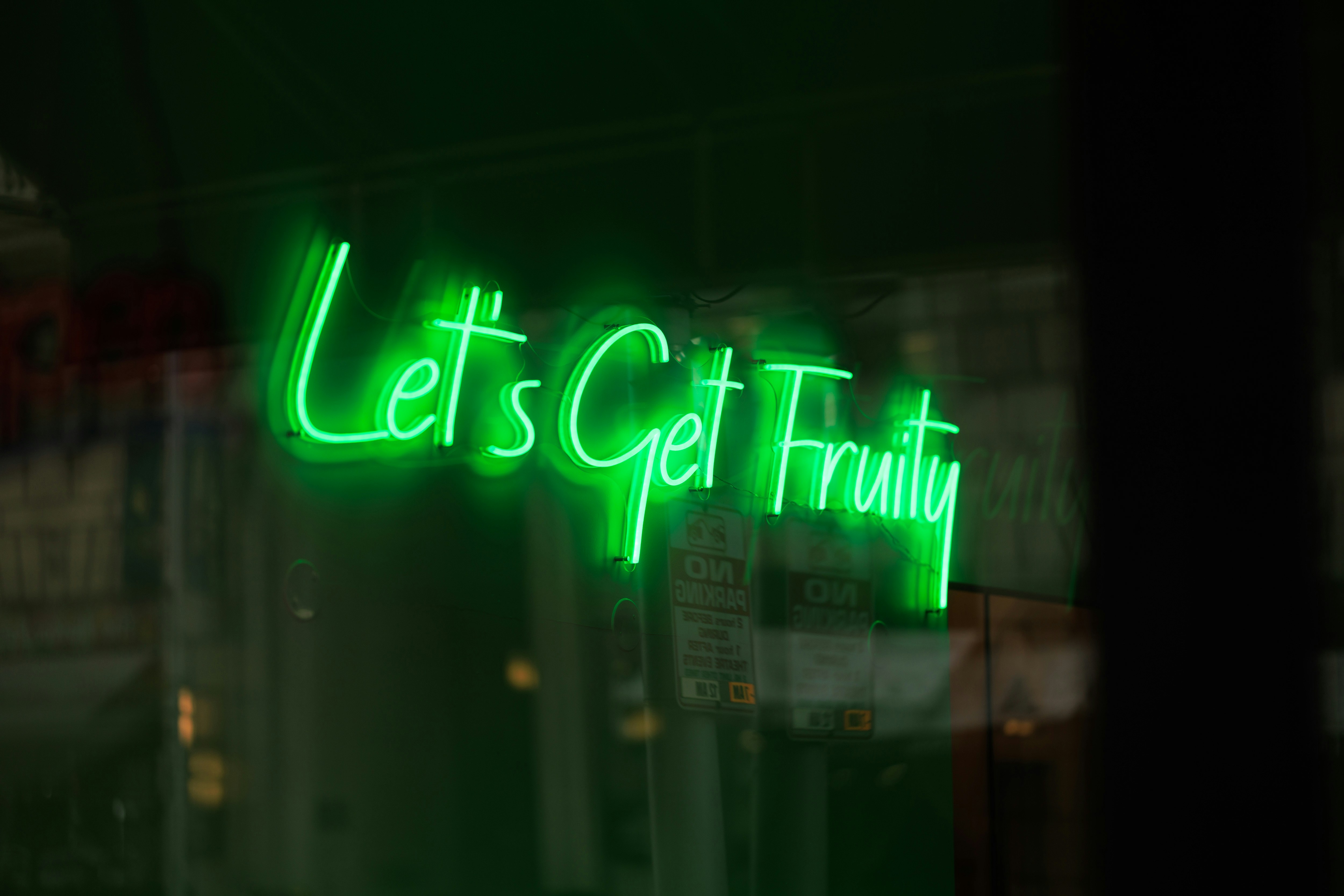 a neon sign in a storefront window that reads "let's get fruity" in cursive, green neon lettering.  the sign is reflecting in the glass.  there are two white paper signs hanging on the inside of the window but the text is not legible. the interior of the store is dark.  the storefront is reflected in the window and appears to be in a city.