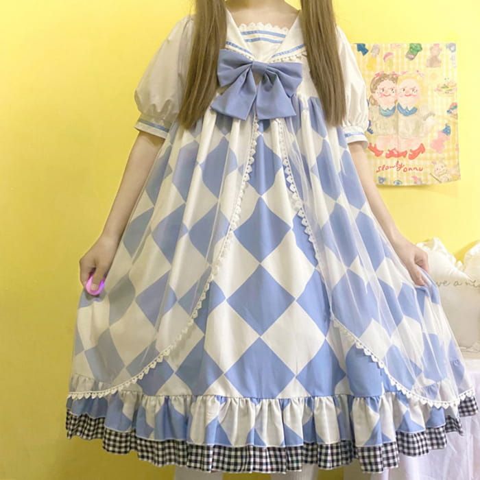 a young woman is wearing a blue and white checkered lolita fashion dress. the dress has a large bow at the neckline and is adorned with lace and frills. the photo is taken indoors in a bedroom with natural lighting.