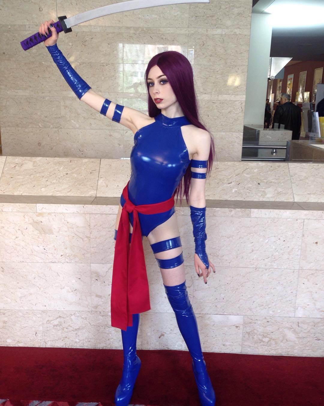 the image shows a young woman cosplaying as psylocke from the xmen comic books and movies. she is dressed in a meticulously crafted blue latex bodysuit with a red sash, and matching thighhigh boots.  she has long purple hair styled similarly to the character. she is holding a katana, and strikes a confident pose in what appears to be an indoor convention setting with marblelike walls and a red carpet floor.  the overall effect is a highly accurate and wellexecuted portrayal of the character.