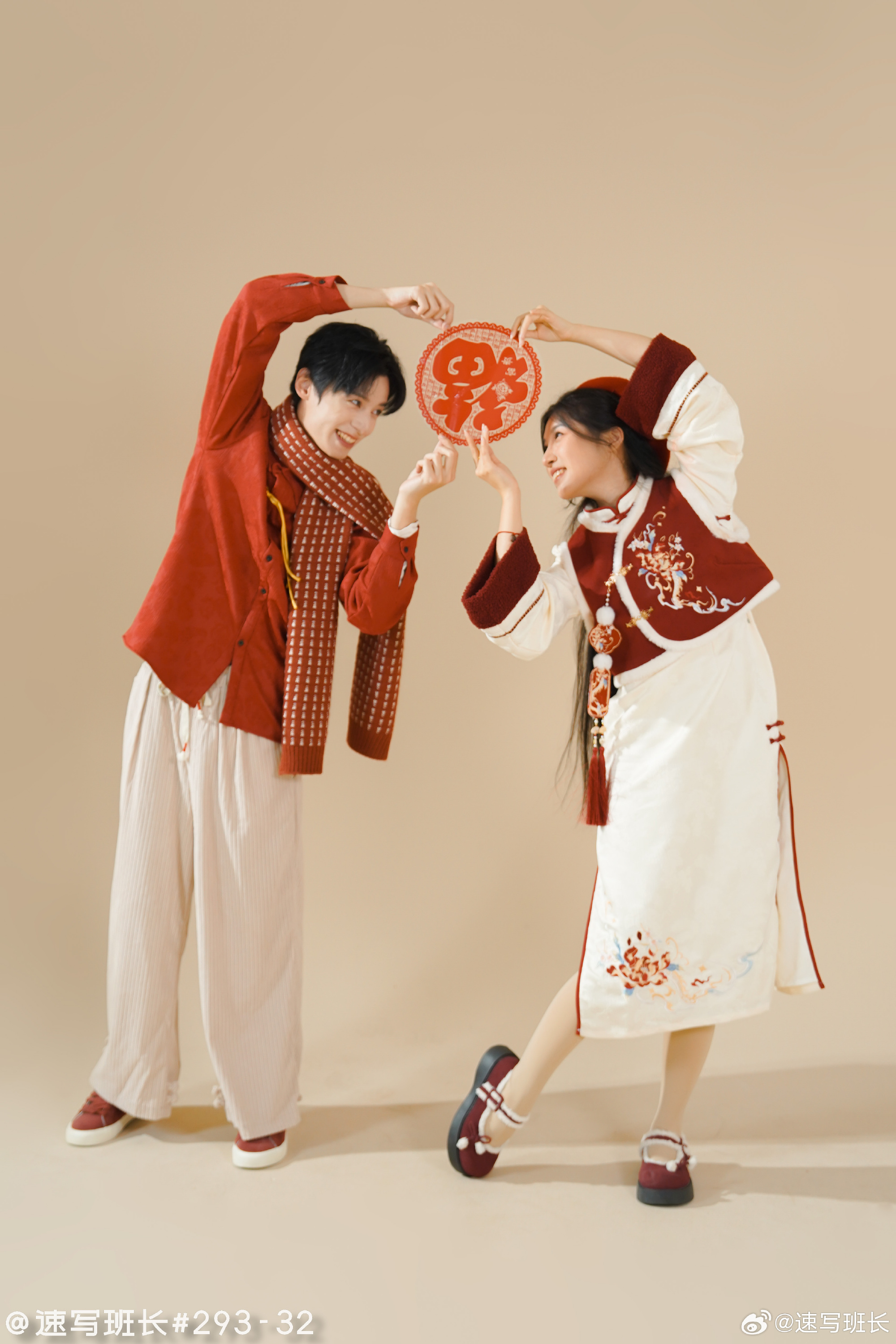 a young man and woman are posing in a studio setting, dressed in traditional chinese clothing for chinese new year. they are both smiling and holding a red paper cutout of the chinese character for "luck". the man is wearing a red jacket, white pants, and a red scarf. the woman is wearing a white dress with red embroidery and a red vest. they are both wearing black shoes. the background is a light beige color.