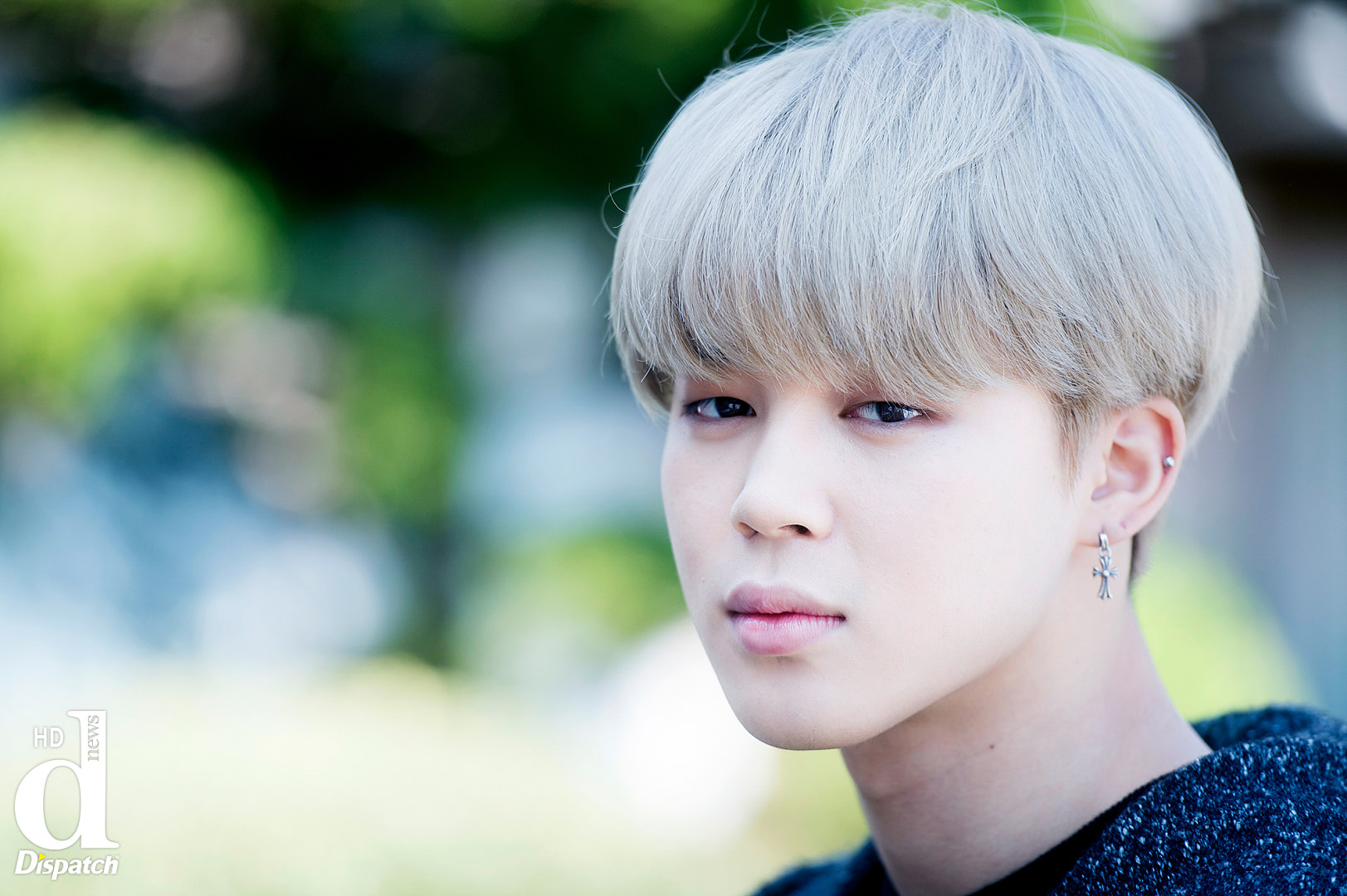 the image is a closeup portrait of a man, identified as jimin, a kpop singer. he has light blonde hair styled in a soft fringe and is wearing a cross earring and an ear piercing. the photo is credited to dispatch.