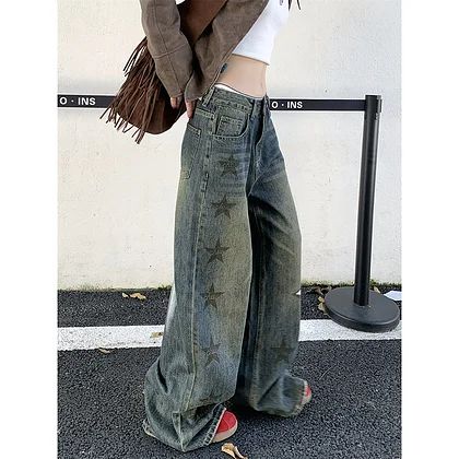 a woman is standing on a pavement next to a barrier wearing a casual outfit consisting of wideleg, highwaisted denim jeans with star patterns, a white top, and a brown jacket. she also has a brown shoulder bag.