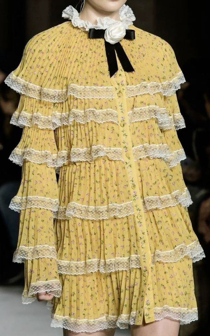 a model wearing a yellow floral dress with ruffled tiers and lace accents on a runway. the dress is pleated and has a vintage feel, giving it a romantic and delicate look. the model is wearing a black bow tie and a white collar, completing the chic and elegant ensemble.